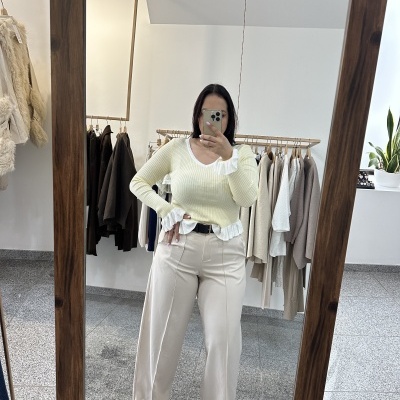 Mulher em espelho com camisola amarela e calças bege numa loja de roupa com cabides ao fundo.