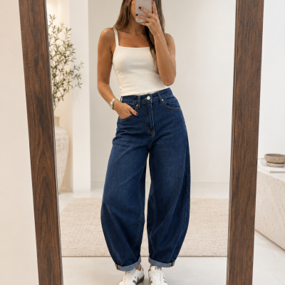Mulher com calças de ganga largas azuis e top branco a tirar selfie em espelho numa sala clara.