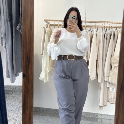 Pessoa com blusa branca e calças cinza, refletida em espelho numa loja de roupa com várias peças penduradas.