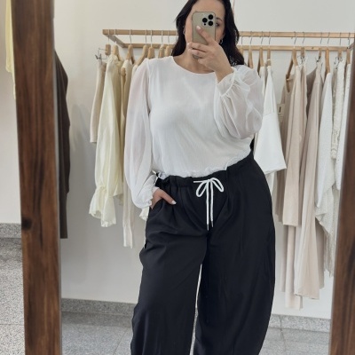 Pessoa com blusa branca de mangas bufantes e calças pretas com cordão branco num espelho, com roupas claras penduradas ao fundo.