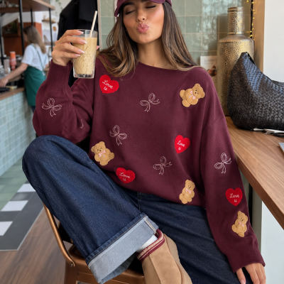 Mulher sentada num café com suéter bordô estampado, calças jeans, boné bordô com texto NEW YORK Est. 1624 e bebida em copo com canudo