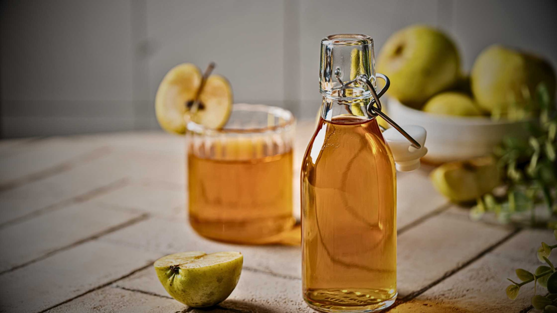 Garrafas e copo com líquido âmbar, maçãs verdes ao fundo, sobre mesa de madeira clara.