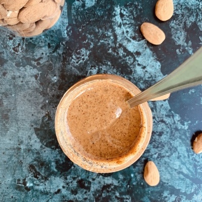 Frasco com creme de amêndoa castanha e colher, amêndoas ao redor, superfície azul escura