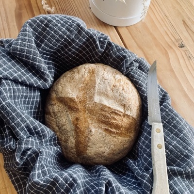 Pão redondo com crosta marcada sobre pano azul com grelha branca ao lado de faca sobre mesa de madeira