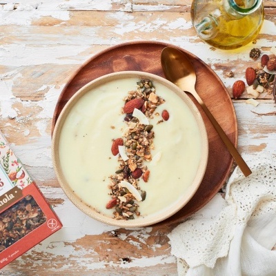 Taça com creme branco e granola, pacote Três Marias Avelã & Coco, colher dourada, mesa de madeira branca