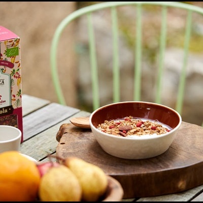 Taça de granola sobre mesa de madeira com caixa e frutas ao lado