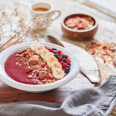 Taça branca com smoothie vermelho decorado com fruta e granola numa mesa de madeira