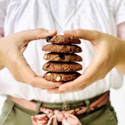 Pessoa segurando pilha de bolachas de chocolate com nozes, vestindo blusa branca e calças verdes com cinto rosa floral