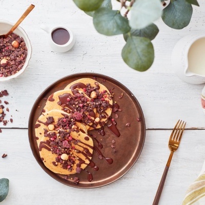 Panquecas com frutas vermelhas e molho num prato castanho numa mesa branca com garfo dourado e jarra de leite