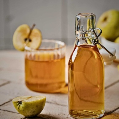 Garrafas e copo com líquido âmbar, maçãs verdes ao fundo, sobre mesa de madeira clara.