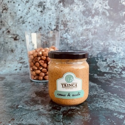 Frasco de creme de avelã TRINCA com vidro de avelãs ao fundo sobre superfície escura