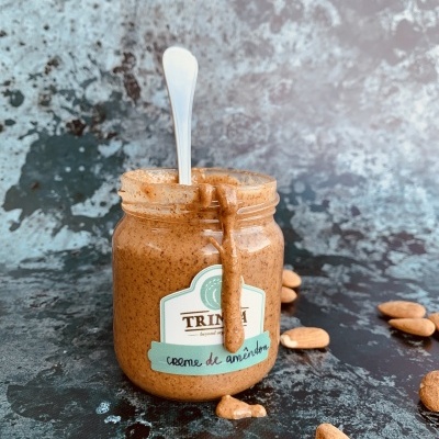 Frasco de vidro com creme de amêndoa castanho e colher, amêndoas inteiras ao redor sobre superfície escura.