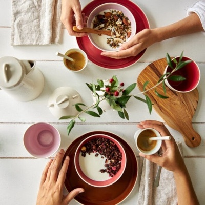 Mesa branca com taças vermelhas com iogurte e cereais, chávenas de café, jarra branca, guardanapos tecidos e ramo decorativo