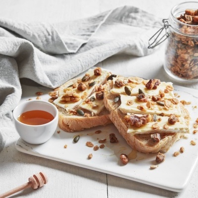 Tostas de pão com banana, granola e mel sobre tábua branca com frasco de granola e pano cinzento