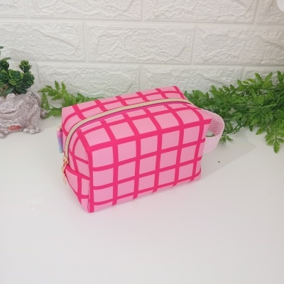 Estuche rectangular rosa con diseño de cuadros y cierre dorado sobre superficie blanca con plantas al fondo.