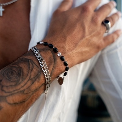 Pulseiras e anel masculinos em metal prateado e contas pretas, com braço tatuado e camisa branca.