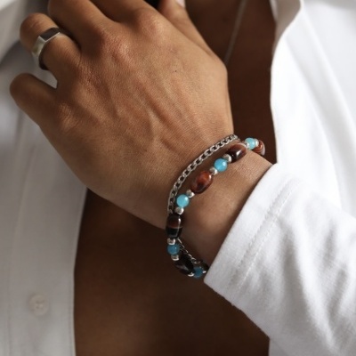 Pulseira masculina com contas azul e castanho e corrente prateada no pulso de homem com camisa branca aberta