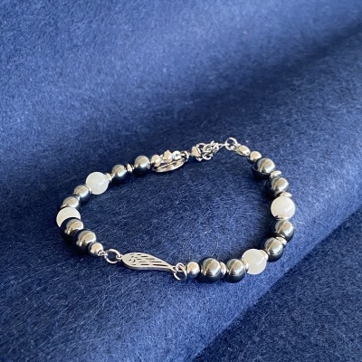 Pulseira de contas prateadas e brancas com pingente em forma de asa sobre tecido azul.