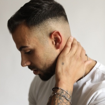 Homem com cabelo curto degradê, tatuagens no braço e t-shirt branca sobre fundo branco.