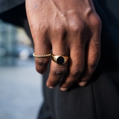 Mão com anéis dourados e pulseiras, roupa preta, ambiente exterior desfocado