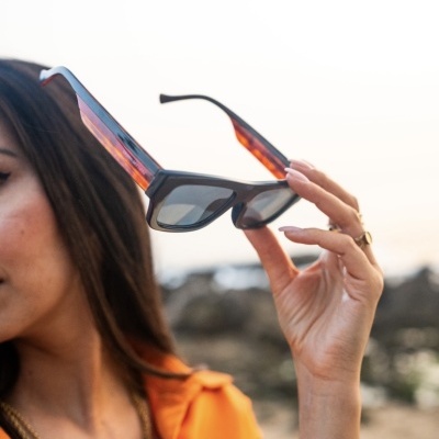 Óculos de sol pretos com hastes castanhas translúcidas a ser segurados por mulher com camisa laranja.