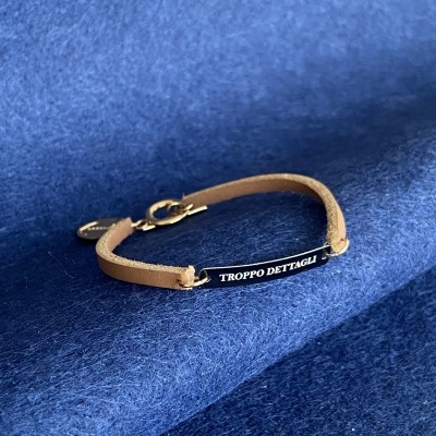 Pulseira de couro bege com placa preta e texto branco TROPP DETTAGLI sobre fundo azul