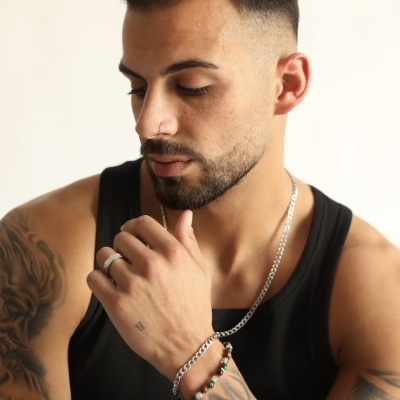 Homem com tatuagens e acessórios de prata, vestindo camisola preta sem mangas.