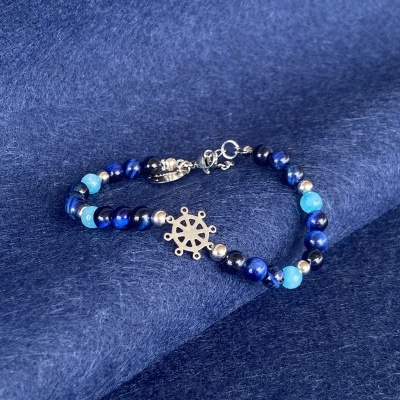 Pulseira de contas azul e prateada com pendente em forma de leme sobre tecido azul.