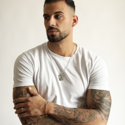 Homem com t-shirt branca, tatuagens nos braços e colar prateado