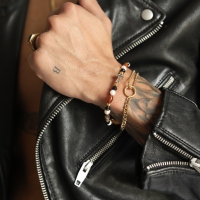 Pulseiras douradas e em contas num pulso com tatuagem, casaco de couro preto com fechos prateados.