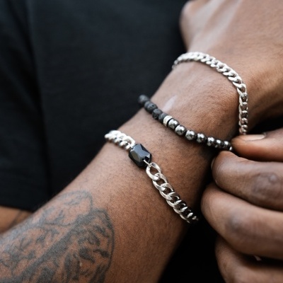 Pulseiras masculinas prateadas e de contas pretas num braço com tatuagem