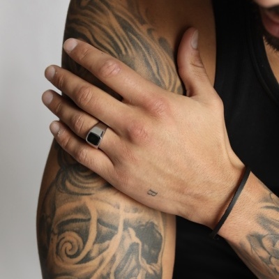 Braço masculino tatuado com anel prateado e pulseira preta em camisa regata preta