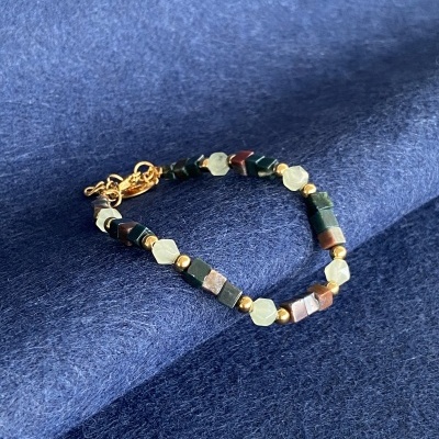 Pulseira com contas hexagonais e cilíndricas verdes, castanhas e bege, com fecho dourado, sobre tecido azul escuro