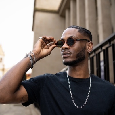 Homem com t-shirt preta, colar prateado e óculos de sol castanhos em ambiente urbano