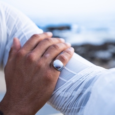 Anel prateado oval num dedo de mão segurando um pulso de camisa branca transparente junto ao mar.