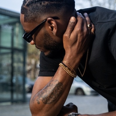 Homem com cabelo trançado e acessórios dourados apoiado num muro de pedra