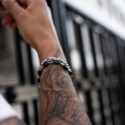 Braço tatuado com pulseira de contas negras e prata junto a uma grade metálica preta