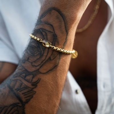 Pulseira dourada de elos largos no pulso de um homem com tatuagem de rosa