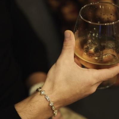 Pulseira dourada com pedras no pulso de homem segurando copo de bebida.