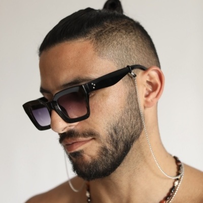 Homem com óculos de sol quadrados e corrente prata, colares de contas, cabelo preso e barba curta.