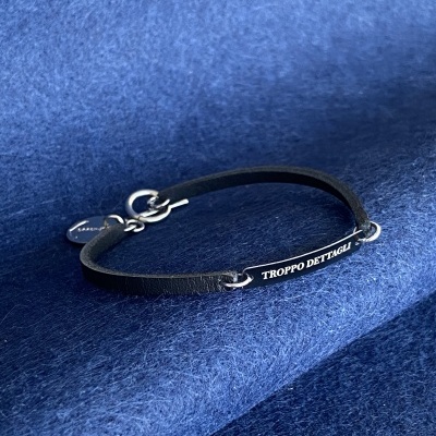 Pulseira preta com placa metálica e texto gravado TROPPPO DETTAGLI em superfície azul.