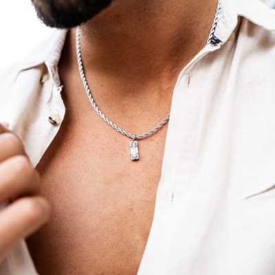 Colar prateado com pendente retangular usado por homem de pele morena com camisa bege aberta