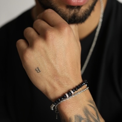 Pulseiras masculinas em contas pretas e prata com tatuagens visíveis no braço e mão, pessoa vestindo t-shirt preta.
