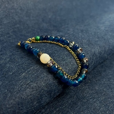 Pulseira com contas azuis, branca, transparentes e verde com corrente dourada sobre fundo azul