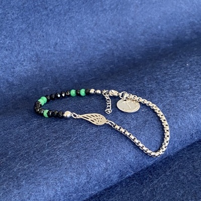 Pulseira mista com contas verdes e pretas e pendentes metálicos sobre tecido azul.