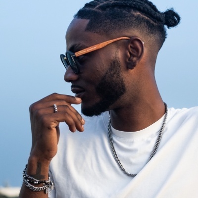 Homem com óculos de sol, cabelo trançado e t-shirt branca ao ar livre.