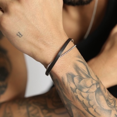 Pulseira masculina de couro preto e corrente prateada no pulso de homem tatuado