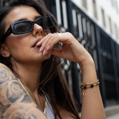 Mulher com óculos de sol pretos retangulares e tatuagem no braço junto a edifício