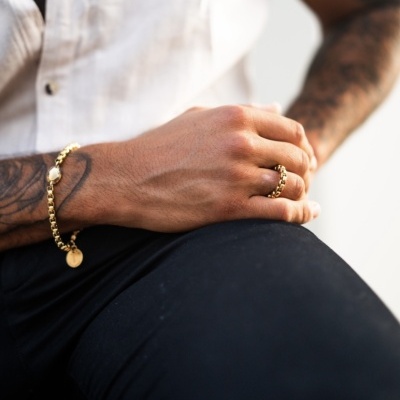 Pulseira e anel dourados com contas redondas em mão direita de pessoa com camisa branca e calças pretas