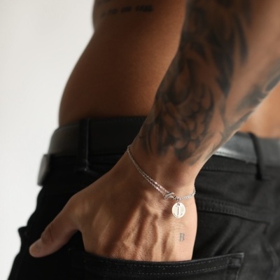 Pulseira prateada com pingente num braço tatuado com mão no bolso de calças pretas
